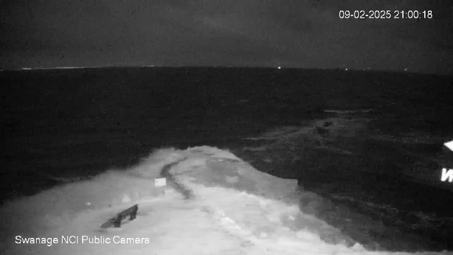 A dark, nighttime scene of the ocean captured by a webcam. The water appears choppy with white foamy waves crashing against a rocky shoreline. In the foreground, there is a small structure visible on the shore, possibly a piece of equipment or debris. The sky is cloudy, and there are faint lights in the distance on the water, likely from boats or ships. The image displays a timestamp, indicating the date and time of the capture.