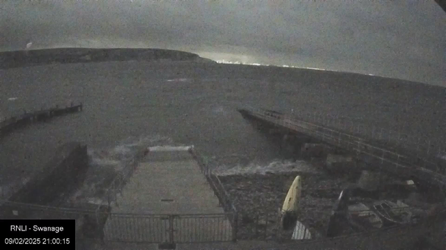 A dark, overcast scene of a coastline at night. In the foreground, there is a rocky area with a concrete platform partially visible. Gentle waves are lapping against the platform. To the right, a small pier extends into the sea with a yellow kayak resting on the ground. The water is grey and choppy, and in the background, the faint outline of a hill and some distant lights can be seen, suggesting a nearby shore or town. The overall atmosphere is calm and slightly eerie due to the lack of light.