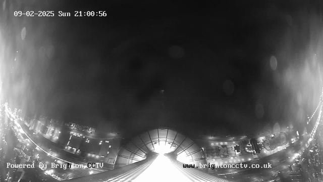 A black-and-white webcam image showing a view of a city at night. The bottom half of the image displays illuminated streets and buildings, with various lights twinkling. The upper portion of the image is dark, with a bright light source at the center, possibly indicating the end of a tunnel or large structure. The date and time are visible at the top, indicating it is Sunday, February 9, 2025, at 21:00:56. The image is branded with "Powered by Brighton CCTV" in the bottom left corner and a web address in the bottom right corner.