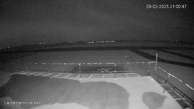 A dimly lit scene shows an empty airfield with a clear patch where a helicopter landing pad is marked. In the background, a faint outline of distant lights can be seen, likely from a nearby town or road. The sky is dark, suggesting nighttime, and there are no visible aircraft or people in the area. A construction barrier is present on the right side, indicating ongoing work or maintenance. The timestamp at the top right shows the date and time.