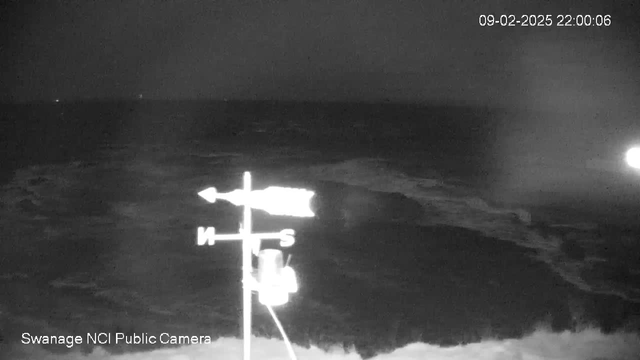 A nighttime image captured by a webcam shows a dark, stormy sea with visible waves. In the foreground, a weather vane points in various directions, with letters indicating cardinal points: North and South. The image is slightly illuminated by an unspecified light source in the background. The date and time of the capture are displayed in the top right corner.