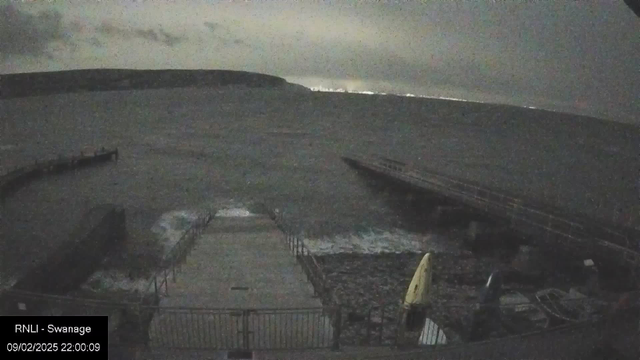 A dark, cloudy night scene at a seaside location. The image shows a partially submerged pier extending into rough waters. Waves are lapping at the base of the pier, and there are several boats tied up nearby. In the background, a faint outline of a coastal hill is visible under the cloudy sky. The atmosphere appears gray and stormy with limited visibility.