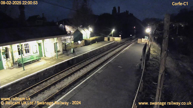 A night view of a railway station platform with stone walls and wooden beams. A green bench is positioned on the left side of the platform. The station building has several windows and is illuminated with warm lights. To the right, a pathway leads out of the main area, marked by a sign that reads "WAY OUT." The two railway tracks run parallel, extending into the distance. There are wooden fences along the platform, and a light post provides additional lighting in the area. The sky is dark, and the surrounding trees are barely visible.