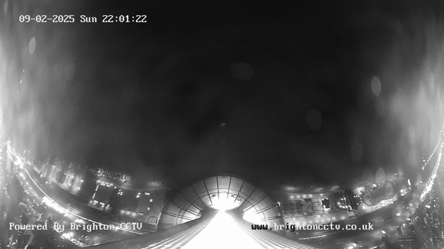 A dark, nighttime scene captured from a webcam, showing a view of a city street illuminated by lights. The image is primarily in black and white, with a bright area in the center suggesting a source of light, likely from buildings or streetlights. The date and time are displayed at the top of the image. The bottom of the image has text indicating it is powered by Brighton CCTV and a website link. The scene appears slightly blurred, possibly due to rain or other atmospheric conditions.