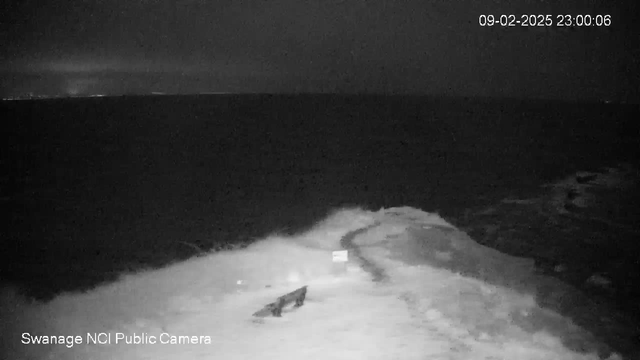 A dark scene showing a nighttime view of a stormy ocean. Waves crash against the rocky shore, with white foam visible. In the foreground, there is a small boat tied to a dock, along with a sign partially obscured by waves. The horizon is faintly lit, suggesting distant lights or buildings. The overall atmosphere is moody and turbulent, typical of a stormy night by the sea.