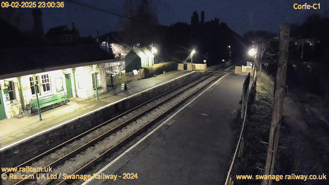 A dimly lit railway station captured at night. The foreground features two tracks running parallel, with a stone platform on either side. To the left, a green bench is situated near a building with large windows that have light emanating from them. Opposite the bench, there is a sign indicating the exit. Further along the platform, a wooden fence delineates the area. The scene is illuminated by streetlights, creating a contrast with the dark surroundings. In the background, faint outlines of trees and buildings can be seen against the night sky.