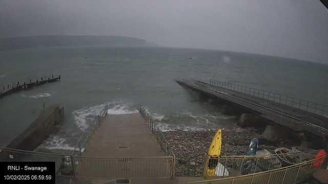 A view of a cloudy marine scene near a pier. The water is choppy with waves crashing onto a rocky shore. Two yellow kayaks and a blue kayak are visible on the right side, resting against the railing near the edge of the water. The pier extends into the water with a platform on the left, and there are steps leading down to the rocky area. The atmosphere appears overcast and moody, suggesting early morning or late evening.