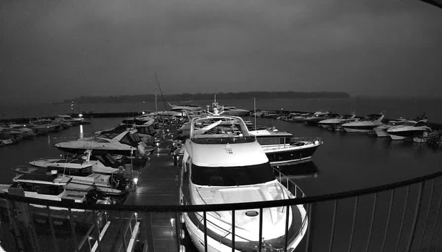 A dimly lit marina filled with various boats docked closely together. The boats are reflected in the still water, creating a mirror-like effect. The sky above is overcast, suggesting a cloudy or stormy atmosphere, with minimal visibility of the landscape in the background. The scene is predominantly black and white, emphasizing the shapes and outlines of the boats and the dock.