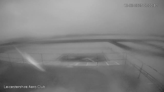 A black and white webcam image showing a blurry scene of an airfield with a runway marked in the center. The image appears misty, with low visibility, and faint outlines of a fence in the foreground. The date and time indicated in the corner is October 2, 2025, at 07:00:13.