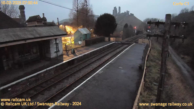 A dimly lit scene at a railway station, with train tracks running through the center. To the left, there is an old stone building with a sloped roof and wooden benches. A small platform area is visible, illuminated by warm lights from a nearby building. In the background, a large hill with ruins can be seen, with several stone towers positioned at the summit. The sky is overcast, and there are electric poles along the right edge, casting a shadow over the tracks.