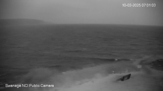 A black and white image of turbulent waves in the ocean under overcast skies. The horizon is barely visible where the water meets the sky. A small boat can be seen partially submerged in the waves near the shore. The scene appears dark and moody, indicating stormy weather. The timestamp at the top right shows the date and time as February 10, 2025, at 7:01 AM. Bottom left text indicates the source as "Swanage NCI Public Camera."