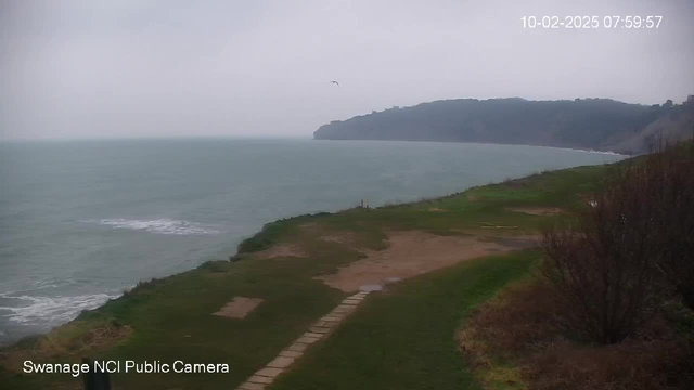 A coastal scene showing a gray, overcast sky above a calm sea. The shoreline is visible with gentle waves approaching the shore. In the foreground, there is a grassy area with a dirt path leading towards the water, bordered by bushes on the right. A small cliff is seen in the distance on the right side of the image. The time and date are displayed in the upper right corner.