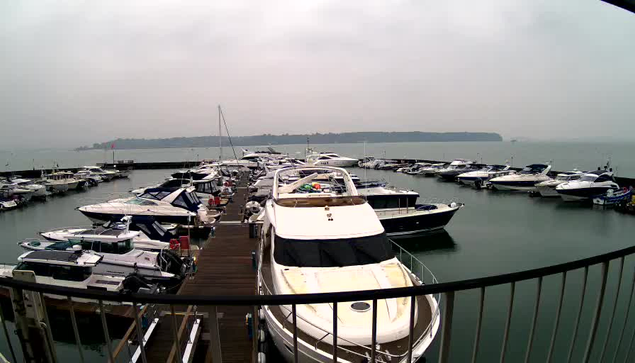 A marina filled with various boats and yachts docked at the pier. The water is calm and reflects the boats. The sky is overcast and gray, indicating a cloudy day. In the background, there is a landmass, likely a shoreline or island, visible on the horizon. A wooden walkway is present in the foreground, leading to the boats.