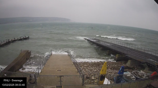 A view of a rocky shoreline with choppy water. There are two piers extending into the sea: one wooden with a railing and another stone structure at the left. Steps lead down to the water from the stone area, which is covered in pebbles. Two kayaks, one yellow and one blue, are on the right side, alongside several piles of equipment. The scene is overcast, with cloudy skies, creating a dull atmosphere.