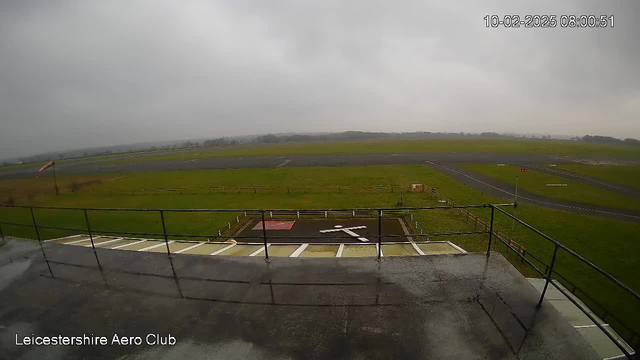 A cloudy sky is visible above an open grass area that serves as an airstrip. The foreground features a railing suggesting an elevated viewpoint at an aero club. Below the railing, there is a red square marked on the ground with a white cross, indicating a helipad or landing area. A portion of the airstrip is seen with a long stretch of asphalt and some fencing in the background. The overall scene is misty, giving it a somewhat muted appearance.