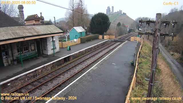 A view of Corfe Castle station in winter. The platform is empty, with two sets of railway tracks running parallel. There is a stone building with a sloped roof and a green waiting bench in the foreground. Behind the building, there is a small green shed. A "WAY OUT" sign is visible on the ground. In the background, Corfe Castle is situated on a hill, partially shrouded in fog. There are trees and a power pole near the tracks, with signs of a rural setting.