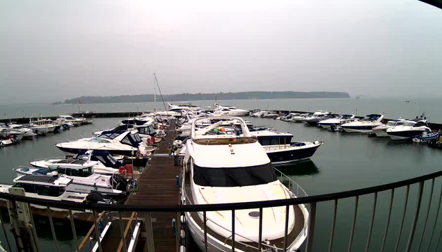 A view of a marina with multiple boats docked. The water is calm and reflects the gray sky above, which indicates an overcast day. In the foreground, several boats, including a large white yacht and smaller vessels, are moored next to a wooden pier. The distant shore is visible on the horizon, with trees and possibly buildings silhouetted against the sky. The scene conveys a tranquil, but slightly gloomy atmosphere due to the lack of sunlight.