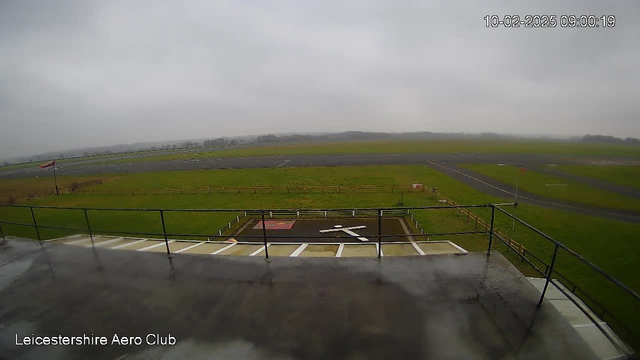 A view from a webcam overlooking an airport runway. The sky is overcast and gray, indicating cloudy weather. In the foreground, there is a railing and part of a roof visible. Below, a grassy area and a runway can be seen, with a large white 'X' marking on a flat surface which is likely a landing zone for aircraft. There is also a red and white windsock on a pole, indicating wind direction. The background is blurred, showing more grass and possibly trees in the distance. The timestamp indicates it is early morning.