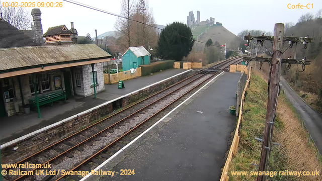 A railway station platform is visible in the foreground, featuring a stone building with a sloped roof on the left. There are benches and signage. In the background, railway tracks stretch into the distance, leading towards a green hillside topped with ruins. A wooden fence separates the platform from the adjacent area, and a telephone pole with wires stands to the right. The sky is overcast, creating a gray atmosphere.