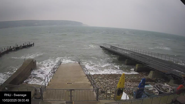 A cloudy scene of a coastal area with a choppy sea. In the foreground, there are concrete steps leading down to the water, bordered by a railing. To the left, there is a small rocky area, while a wooden jetty extends into the water on the right side. Several kayaks are stored near the jetty, with one yellow kayak visible. The landscape in the background features a hillside that slopes gently toward the sea. The overall atmosphere appears misty and overcast.