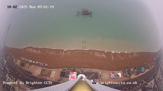 Aerial view overlooking a sandy beach and the sea. The water is a light blue, with a few scattered wooden posts visible in the surf. There is an old pier structure partially submerged in the water. The beach is lined with a promenade featuring buildings and attractions, including a circular structure and a colored water feature. The image is timestamped at 09:01 on February 10, 2025, and displays a gray sky, likely indicating cloudy weather.