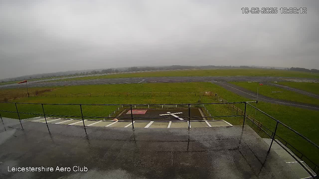 A view from a webcam showing a gray, overcast sky. In the foreground, there is a balcony railing. Below, a grassy area is visible with a helipad marked by a white cross on a red square. Beyond the helipad, there is a small strip of tarmac marked for landing, and further back, a larger runway. Surrounding the grass area is a wooden fence. The scene appears wet, indicating recent rain.