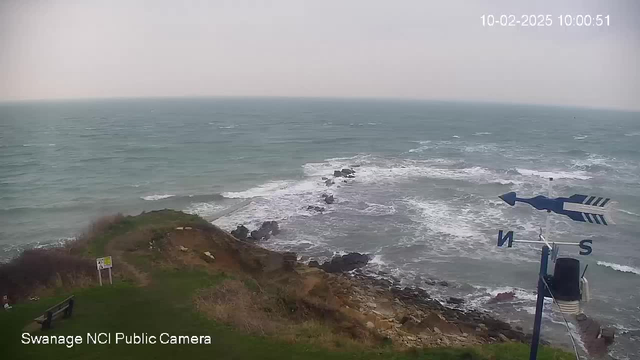 A view of the ocean taken from a cliffside. The water is a dull blue-gray, with waves creating white foam as they crash against the rocks below. In the foreground, there's a grassy area with a few rocks, and a sign on a post is partially visible. To the right, a weather vane is shown, indicating wind direction with the cardinal points marked. The sky appears cloudy and overcast.