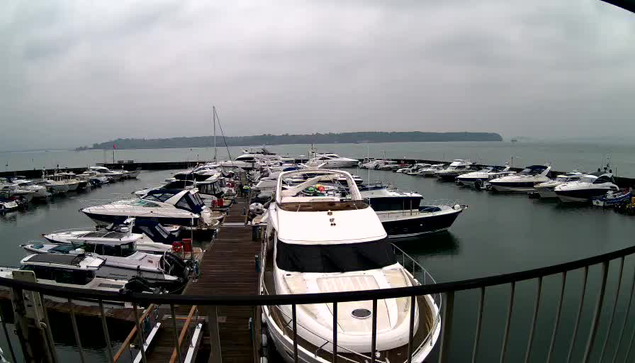 A view of a marina on a cloudy day. Numerous boats are docked on calm waters. In the foreground, a large white motorboat is prominent, with several smaller boats surrounding it. The background features a hazy line of land or hills in the distance, partially obscured by clouds. The scene conveys a quiet, serene atmosphere.