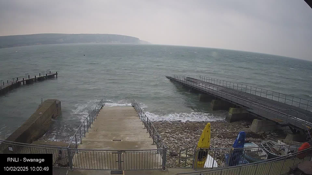 A coastal scene showing a staircase leading down to a rocky beach with crashing waves. In the foreground, there are two kayaks, one yellow and one blue. On the left, a stone wall is visible, and beyond it, there are two wooden piers extending into the water. The sky is overcast, and distant cliffs are visible in the background.