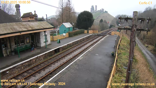 The image shows a train station scene with a platform on the left side. There is a green bench and a signpost nearby. A person wearing a black coat walks across the platform. In the background, there is a low stone wall and a set of train tracks leading away. A small turquoise building is visible, along with a wooden fence that encloses a grassy area. On a hill in the distance, the ruins of a castle can be seen. The sky is overcast, giving the scene a muted appearance.