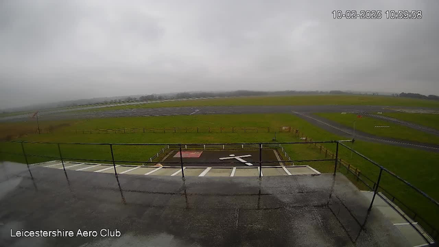 A cloudy scene from a webcam overlooking a large open field. In the foreground, there is a railing and a helipad marked with a white cross. The ground is wet, likely from recent rain. In the background, there is an airport runway with a grassy area and some trees in the distance. The visibility appears limited due to overcast skies.