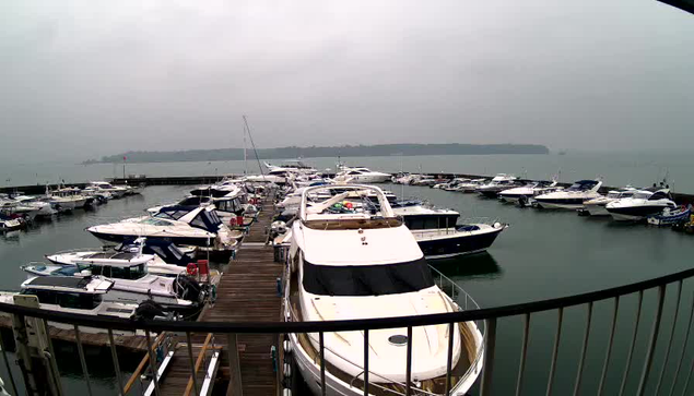 A view from a webcam showing a marina filled with various boats docked in calm water. In the foreground, there is a large white yacht with a covered cockpit. Multiple other boats, including smaller motorboats and sailboats, are visible throughout the marina. The sky is overcast and gray, suggesting a cloudy day. The image captures the stillness of the water and the arrangement of the boats in the harbor.