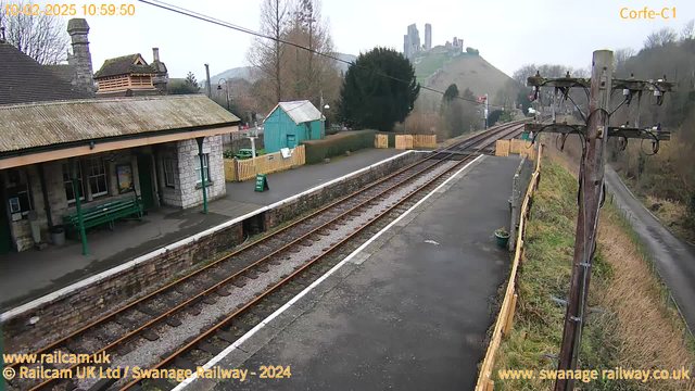 A view of a train station platform with wooden benches under a covered area. The platform features brown railway tracks and a concrete walkway. On the left, there is a stone building with a sloped roof, and on the right, a green wooden shed. There is a sign on the platform that reads "WAY OUT." In the background, there are ruins of what appears to be a castle situated on a hill. The sky is overcast, and there are trees lining the area. A wooden pole with electrical wires is located on the far right side.