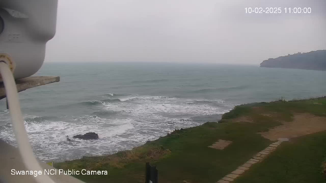 A cloudy view of the sea where gentle waves roll onto a rocky shoreline. The foreground features a grassy area, with pathways leading towards the water. A faint outline of a landmass rises in the distance. The image includes a portion of a webcam structure on the left side. The timestamp shows the date as October 2, 2025, and the time as 11:00 AM.