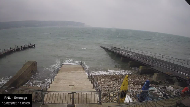 A coastal scene on a cloudy day. In the foreground, a wide concrete ramp leads down to the water, bordered by a metal railing. To the left, a rocky shore is visible. Several colorful kayaks—yellow, blue, and red—are stored on the ground nearby. In the background, the sea is choppy with small waves, and several piers extend out into the water, with low visibility of distant cliffs on the horizon. The overall atmosphere is gray and overcast, suggesting windy weather.