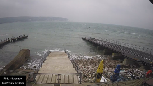 A coastal scene with choppy waters and gray skies. In the foreground, a set of concrete steps leads down to the water, flanked by metal railings. To the left, there is a stone barrier, while on the right, a wooden pier extends into the sea. Several colorful kayaks are stacked near the steps, with a yellow one prominently displayed. The rocky shore can be seen beneath the pier. The overall atmosphere is cloudy and rugged, characteristic of a coastal environment.