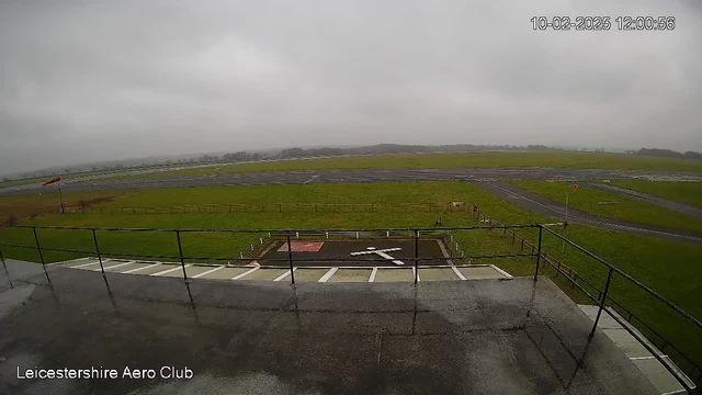 A view from the Leicestershire Aero Club webcam shows a large, slightly wet, flat area with a grassy field and a runway in the background. The sky is overcast and gray, indicating cloudy weather. In the foreground, a small helipad with a white cross is visible, surrounded by a rail fence. A windsock is positioned on the left side of the image, indicating the direction of the wind, and a segment of a runway appears to the right. The overall scene conveys a quiet, open-air environment typically associated with aviation settings.