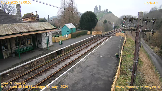 A view of a train station in a rural setting. The foreground features railway tracks with gravel and a small platform made of stone and grass. On the left, there is a stone building with a green roof, and a wooden shelter with benches under the awning. A green shed is visible in the background alongside a low wooden fence. In the distance, a hilly landscape rises, topped with the ruins of a castle. The scene is overcast with muted colors. Utility poles and wires are positioned along the right side of the image.