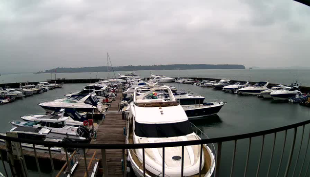 A marina scene under overcast skies, featuring numerous boats docked in calm waters. In the foreground, a large white boat is partially visible, with its cabin and deck facing the camera. Surrounding it are several smaller boats of various sizes and colors, including blue and white. The background shows more boats on the water, with land visible in the distance. The scene conveys a tranquil, nautical atmosphere.