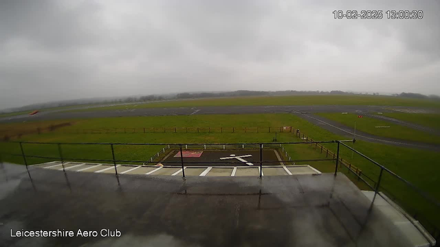 A view from a webcam at Leicestershire Aero Club shows a grassy airfield under cloudy, overcast skies. In the foreground, there is a rooftop balcony or platform with a railing. Below, a helipad marked by a red square with a white cross is visible. The airfield extends into the distance with several taxiways and grassy areas, while wooden fencing borders parts of the field. The scene is dull and gray due to the cloudy weather.