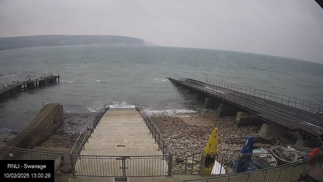 A foggy coastline scene showing a rocky beach with steps leading down to the water. There are two piers extending into the sea on either side of the image. The water appears choppy, and there are a few small boats in the distance. Several colorful kayaks are stacked on the beach near the bottom right corner. The sky is overcast, suggesting a gloomy day.