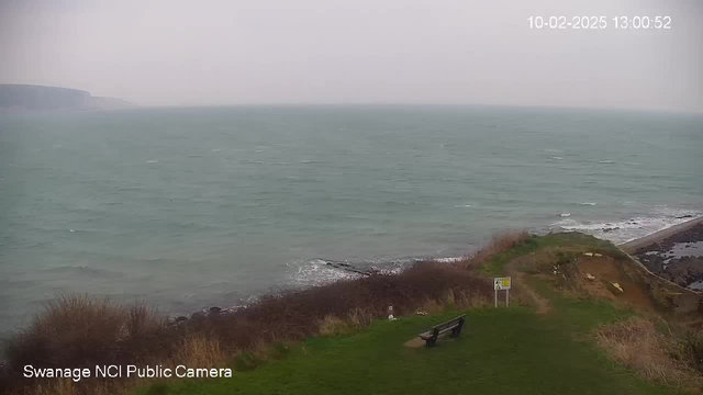 A view of the sea on a cloudy day, with choppy waves gently rolling towards the shore. In the foreground, there is a grassy area with a wooden bench facing the water. To the right, a sign is partially visible, and to the left, there are coastal cliffs that fade into the mist. The sky is gray and overcast, with no discernible sunlight.