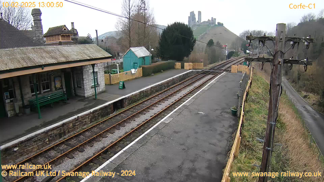 A view of a train station with a stone building featuring a sloped roof on the left. There is a green bench under a covered area. The station platform has visible railway tracks extending into the distance on the right side. In the background, hills rise with a castle ruin perched on top, surrounded by trees. A green wooden shed is located between the station building and the tracks, and there is a sign reading "WAY OUT" visible on the platform. The scene is set during a cloudy day, with no trains present on the tracks.