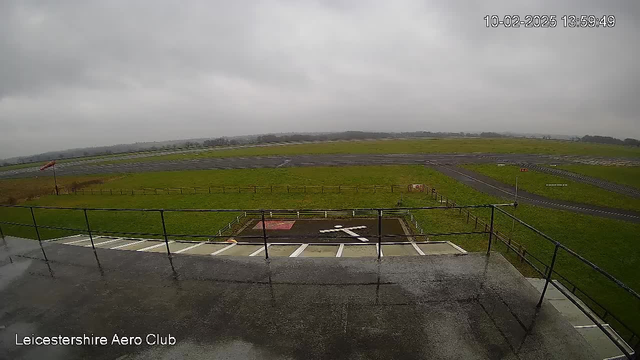 A cloudy sky is visible above a grassy field. In the foreground, there is a viewing platform with railing. Below the platform, a white cross is marked on a dark surface, likely indicating a landing area. In the background, a runway with a few scattered markings is seen, along with a fence framing the grassy area. The overall scene appears overcast and damp.