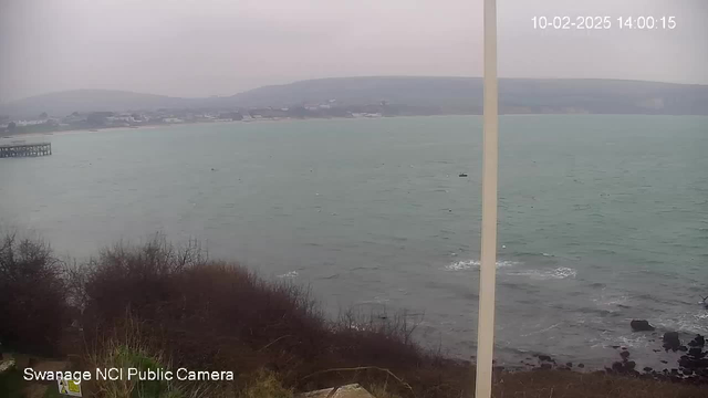 A cloudy day overlooking a calm turquoise sea, with gentle waves breaking at the shoreline. In the distance, there is a wooden pier extending into the water. The coastline is lined with low hills and sparse buildings, partially obscured by mist. In the foreground, there are patches of dry grass and rocks. A camera frame and timestamp are visible in the corner.