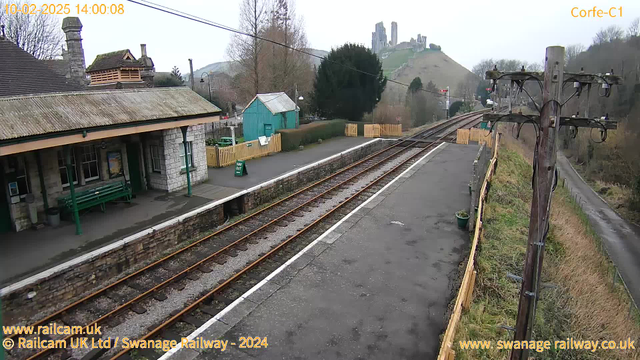 An outdoor view of a railway station with a stone building featuring a sloped roof and a covered waiting area with benches. In the background, a small green shed is visible, along with a wooden fence outlined by a path. The railway tracks run alongside the platform, leading into the distance. On a hill behind the station, there are ruins of a castle. The sky is overcast with limited visibility, and a wooden telephone pole stands on the right side of the image.