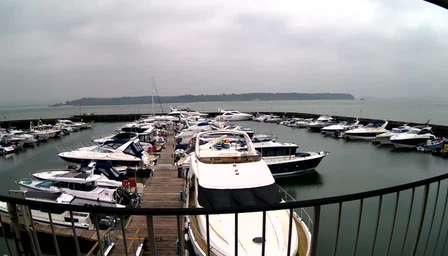 A view of a marina on a cloudy day, featuring multiple boats docked in calm water. The boats are varied in size and type, with some covered and others exposed. Wooden docks extend between the boats, and a distant shoreline is visible in the background. The sky is overcast, contributing to a gray atmosphere.