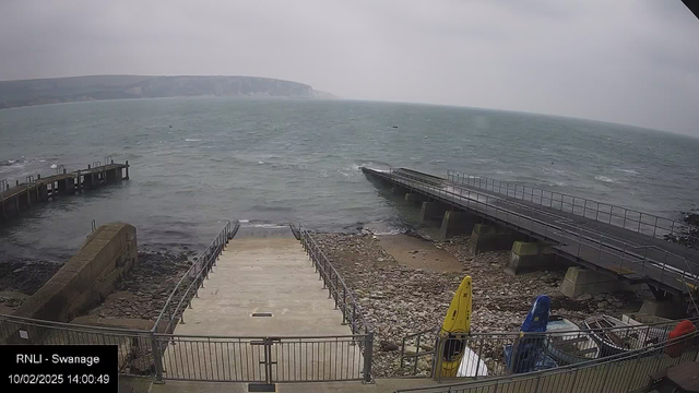 A cloudy day at a coastal area, with a view of the sea and rocky shore. In the foreground, a set of stairs leads down to the water, flanked by a railing. To the right, there is a small dock extending into the water, with several boats visible. The water is choppy, and a distant cliff can be seen in the background. The image has a timestamp indicating the date and time.