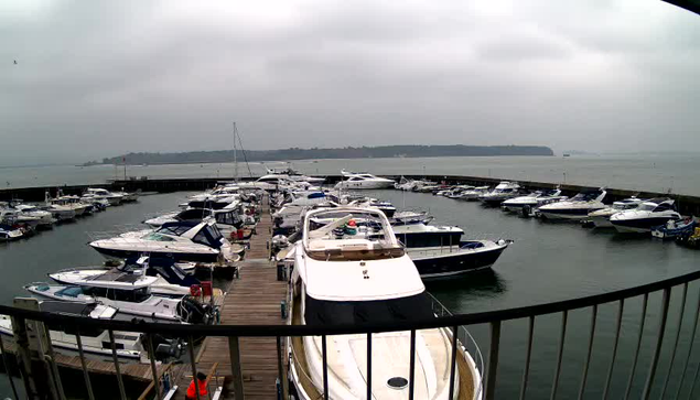 A cloudy day at a marina with numerous boats docked in the water. The scene includes various types of boats, some larger motor yachts and smaller vessels, arranged along a wooden pier. Water ripples reflect the gray sky, and a distant landmass can be seen in the background. The marina is surrounded by a low railing, and there are hints of colorful equipment and safety markers on the docks.