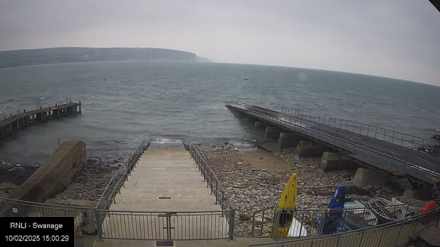 A cloudy day at a coastal location with gray skies. The image shows a view of the sea with gentle waves. In the foreground, there are steps leading down to the water, flanked by a railing. To the left, there's a small pier extending into the water. Nearby, several colorful kayaks in yellow, blue, and red are positioned on the shore. Rocky and pebbly areas are visible along the water's edge. In the background, green hills rise along the coastline.